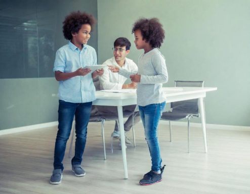 Dos niños conversan en un ambiente interior moderno, con un adulto al fondo.
